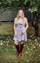 Load image into Gallery viewer, Blonde hair model in a light blue wrap dress with a small print. Model is standing in a garden under an apple tree.