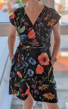 Load image into Gallery viewer, Picture of a black wrap dress with big floral print in red, blue, green and yellow. Model has hands in the side pockets. No head, just the dress.